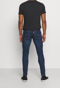 Vaqueros ajustados de color azul oscuro con bolsillos traseros, combinados con una camiseta negra de cuello redondo y zapatillas negras con suelas blancas.