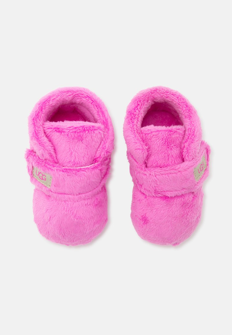 Chaussons pour bébé doux et pelucheux en rose vif avec des brides ajustables et des patchs avec le logo UGG sur un fond uni.