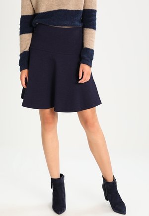 Kobieta ubrana w granatową rozkloszowaną spódnicę, beżowo-granatowy sweter w paski i granatowe zamszowe botki stojąca na jednolitym tle.