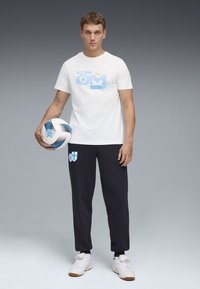 Wit T-shirt met blauwe grafische print, zwarte trainingsbroek met logo, een voetbal met patroon vasthoudend, op een grijze verloopachtergrond.