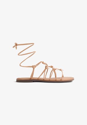 Sandalo piatto color sabbia con molteplici sottili cinturini, dettagli perlati e un laccio alla caviglia con un piccolo ciondolo dorato su sfondo bianco.