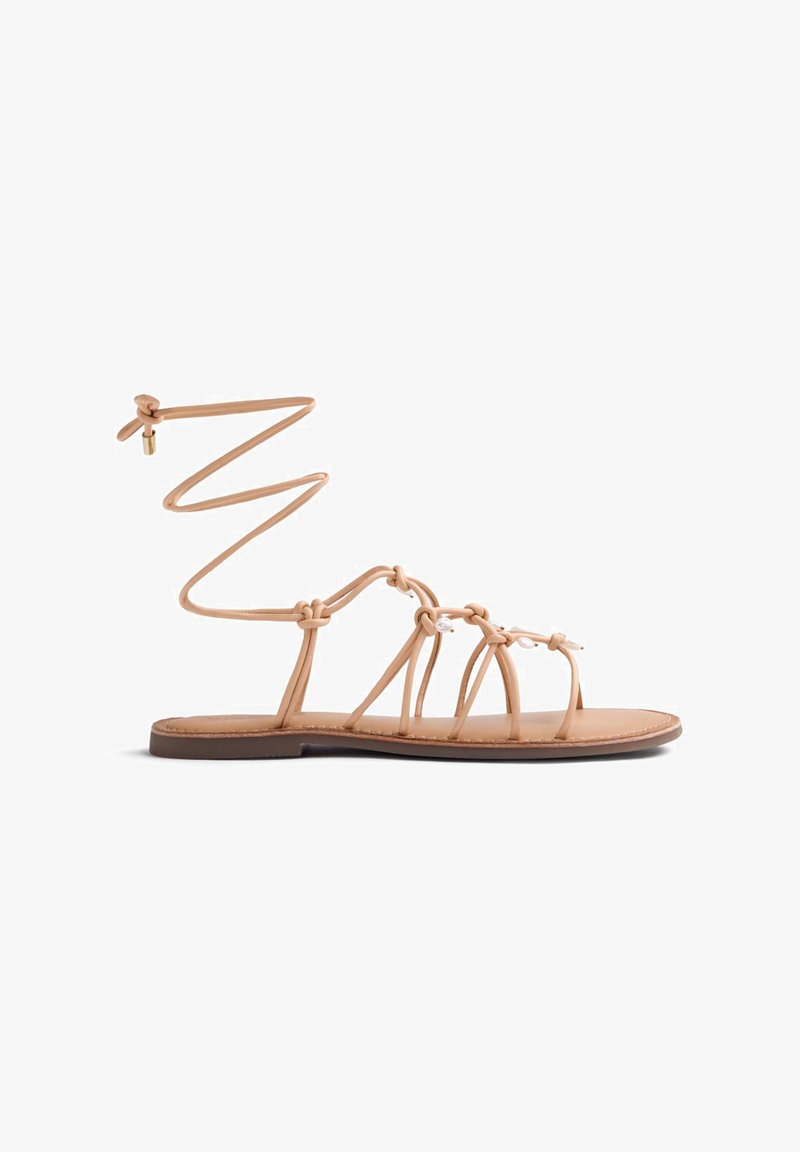 Sandale plate beige avec plusieurs fines brides, accents de perles et un lien à nouer à la cheville orné d'un petit charme doré sur fond blanc.