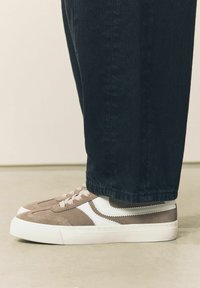 Brune og hvide sneakers har suede og lædermaterialer, en tekstureret overflade, rund tå og hvid gummisål, båret med mørke denimbukser.