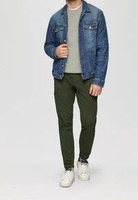 Homme portant une veste en denim bleu, un sweat-shirt gris clair, un pantalon jogger cargo vert olive et des baskets blanches, debout devant un fond uni.