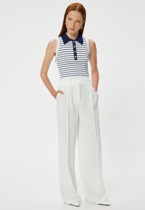 Kvinde med rødt hår iført en ærmeløs hvid og navystribet strikket top og hvide, høj taljede bukser med vide ben, hænderne i lommerne.