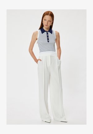 Frau mit roten Haaren, die ein ärmelloses weißes und marineblau gestreiftes Stricktop und weiße, hoch taillierte, weit geschnittene Hosen trägt, Hände in den Taschen.