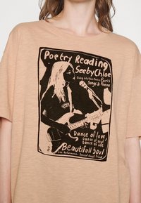 Beżowy bawełniany t-shirt z czarnym nadrukiem graficznym przedstawiającym kobietę z gitarą, tekst o wieczorze poezji oraz w stylu vintage.