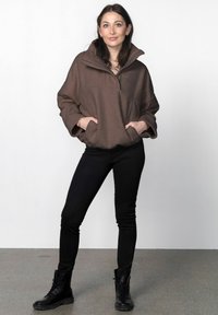 Hnedý fleece pulóver s vysokým golierom a veľkými vreckami, nosený s čiernymi úzkymi nohavicami a čiernymi šnurovacími čižmami, na bielom pozadí.