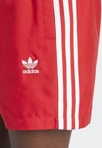 Κόκκινα αθλητικά σορτς από λείο ύφασμα, με τρεις λευκές ρίγες στα πλάγια και ένα λευκό κεντημένο λογότυπο στο κάτω μέρος.