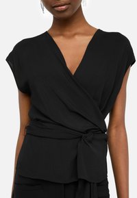Top avvolgente nero realizzato in tessuto leggero, con scollatura a V profonda, maniche a sbuffo e dettaglio di cintura annodata in vita.