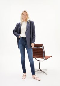 Blazer azul marino, suéter crema, jeans azules ajustados y zapatos rosas. Diseño moderno con textura suave y estilo minimalista. Silla marrón en el fondo.