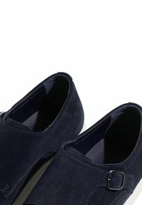 Mornarsko plave suprotne cipele s ravnim bijelim potplatom, s detaljem kopče sa strane i šivenim akcentima, ističući glatku teksturu.