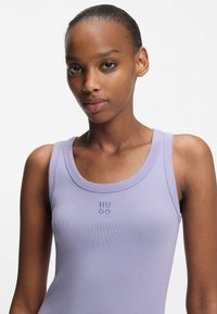 Laventelin värinen hihaton toppi pyöreällä pääntiellä, jossa rinnassa kirjailtu teksti "HU GO" kontrastivärillä. Sileä kangas.