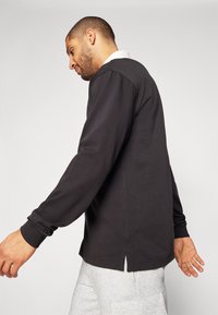 Črna dolga majica z dolgimi rokavi in belim ovratnikom, ki ima sproščen kroj in stranske razpone na robu. Iz bombažnega materiala z gladko teksturo.