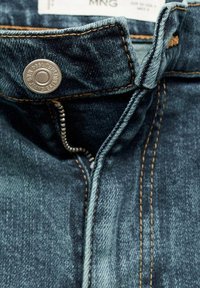 Gros plan d’un jean en denim bleu foncé avec une fermeture éclair partiellement ouverte, un bouton en métal et des surpiqûres orange visibles près de la ceinture.