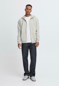 Light gray zip-up hoodie with a drawstring hood, worn over a white t-shirt, paired with dark jeans and gray sneakers. Smooth texture.