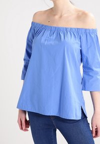 Blusa blu chiaro a spalla scoperta con maniche a tre quarti, collo arricciato e fessure laterali, realizzata in tessuto liscio.