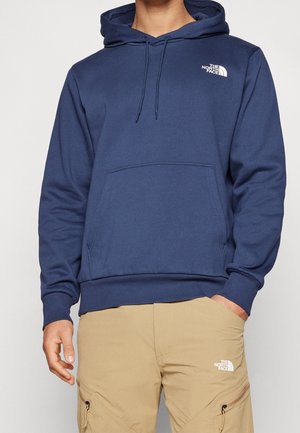 Muž oblečený v tmavě modré mikině The North Face a béžových cargo kalhotách The North Face, stojící s jednou rukou v kapse.