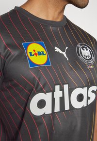 Črn športni dres z rdečimi in oranžnimi diagonalnimi črtami, ki vsebuje veliko belo besedilo "atlas," logotip Puma in znak Lidl.