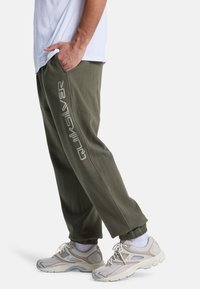 Olivgröna joggarbyxor med elastiska cuffs och sidofickor, med vitt "Quiksilver"-logotryck längs vänster ben. Grå sportskor.