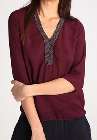 Blouse bordeaux avec un décolleté en V et un col ornés de perles. Dispose de manches trois-quarts et d'un ourlet légèrement froncé. Texture de tissu lisse.