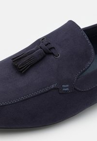 Mocassin en suede bleu foncé avec des tassels décoratifs et des coutures visibles sur le dessus, posé sur un fond uni clair.