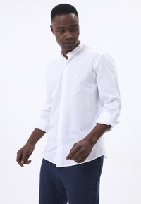 Camisa blanca de manga larga con cuello abotonado, que presenta una sutil textura y un pequeño emblema en el lado izquierdo del pecho. Combinada con pantalones azul oscuro.