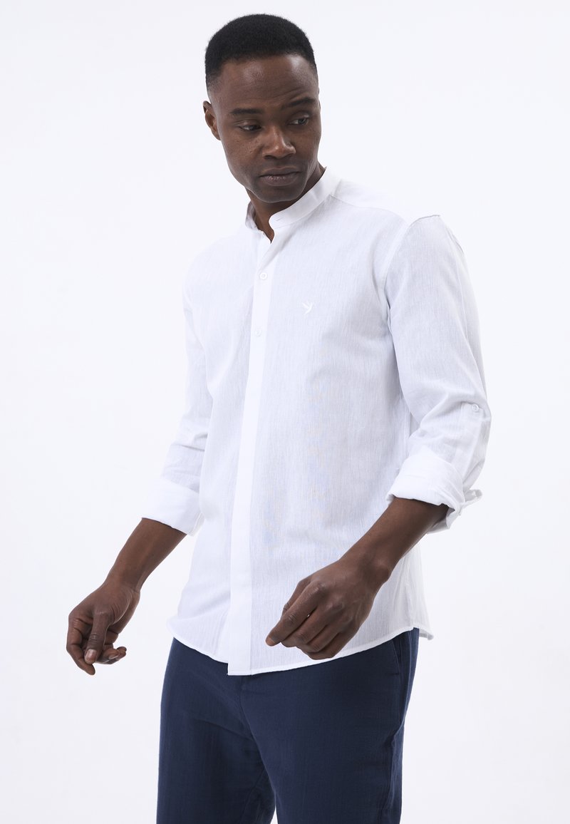 Camisa blanca de manga larga con cuello abotonado, que presenta una sutil textura y un pequeño emblema en el lado izquierdo del pecho. Combinada con pantalones azul oscuro.