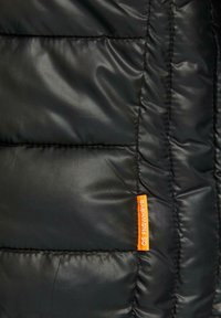 Veste matelassée noire avec texture matelassée, dotée d'une étiquette de marque orange vif. Le matériau semble brillant et résistant à l'eau.