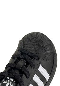 Čierna atletická obuv s textúrovaným gumovým špičkovým krytom, čiernymi šnúrkami a tromi bielymi pruhmi po stranách. Má vzorovaný dizajn na špičke.