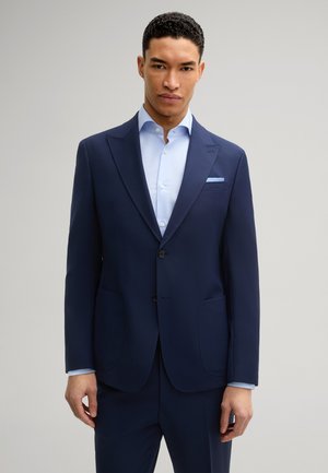 Marineblauwe blazer van soepel materiaal, met twee voorzakken, notch revers en een lichtblauw shirt eronder voor contrast.