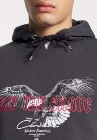 Sudadera con capucha negra y cordón, con un gran gráfico de un águila y el texto "SOLO LOS VALIENTES" en rojo, textura de tela suave.