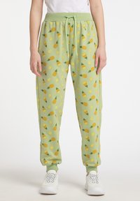 Grüne Jogginghose mit gelben Ananas- und Zitronenmuster, ausgestattet mit einem elastischen Bund, einer Kordel und schmal zulaufenden Bündchen. Getragen mit weißen Sneakern.