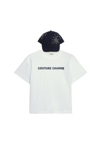 Witte katoenen T-shirt met de tekst "COUTURE CHARME" in marineblauw. Gecombineerd met een marineblauw pet versierd met kleurrijke versieringen.