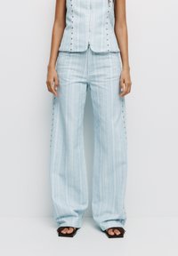 Pantaloni a righe blu chiaro con gambe larghe, dotati di bottoni a pressione argentati lungo le cuciture esterne. Tessuto liscio e design con vestibilità morbida.