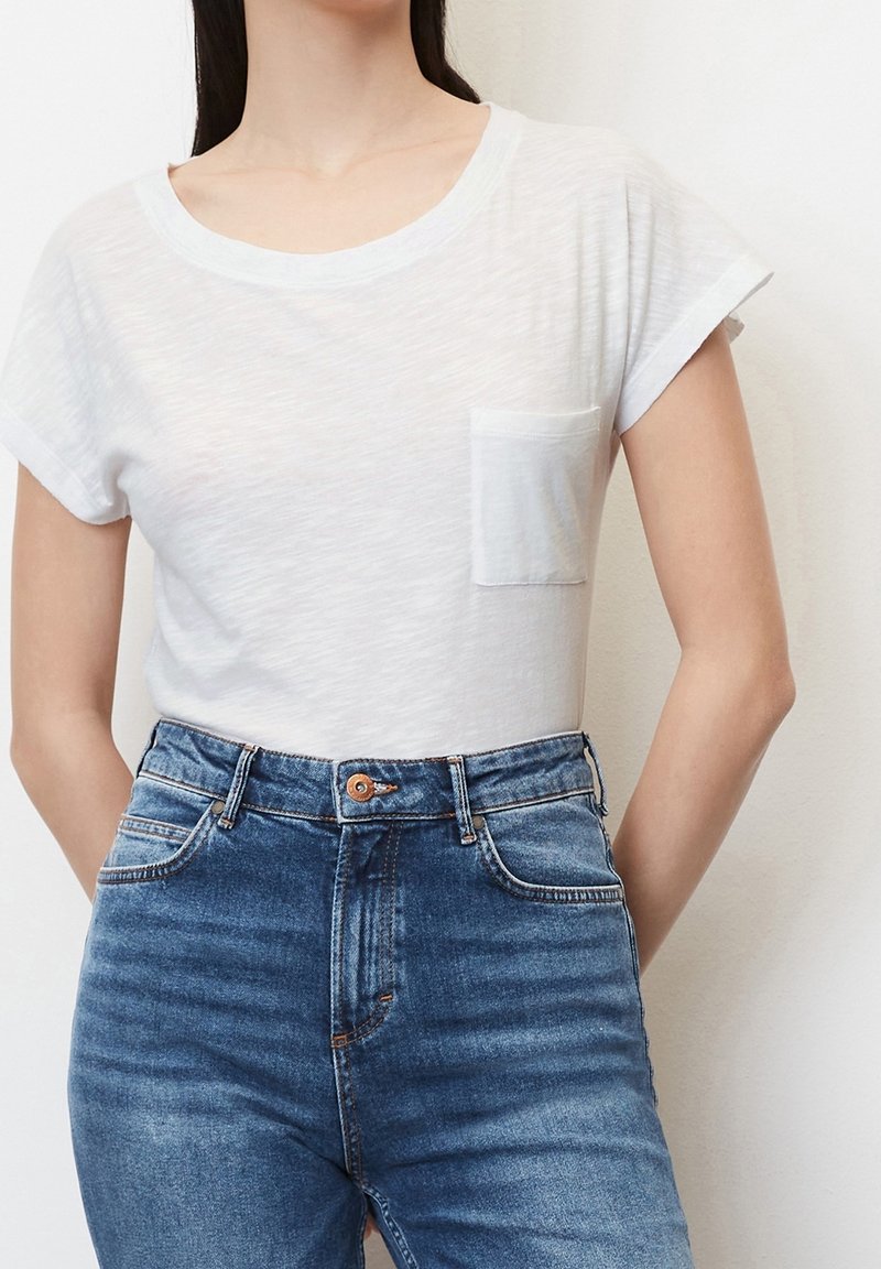 Femme portant un t-shirt blanc à manches courtes avec une poche, associé à un jean bleu taille haute, debout devant un fond uni.
