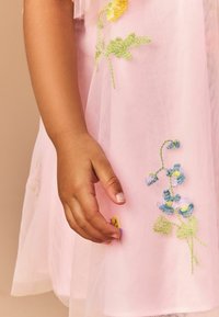 Rosa Tüllkleid mit lebhaften gestickten Blumen in Gelb, Blau und Grün. Verfügt über einen strukturierten, durchsichtigen Stoff. Eine Hand greift nach dem Detail.