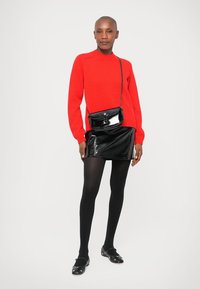 Stoječa oseba, oblečena v rdečo pulover, črn bleščeč mini krilo, črne nogavice do stegen, črne balerinke in majhno črno torbico na čez ramo.