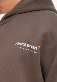 Hnedá mikina s kapucňou a bielym a oranžovým logom tímu McLaren Formula 1 na hornej časti hrudníka blízko ramena.