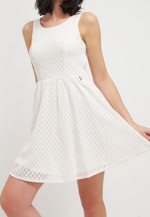 Kobieta w białej, bezrękawnikowej koronkowej sukience z dopasowaną górą i rozkloszowanym dołem, trzymająca sukienkę na bok na jednolitym tle.