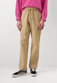 Beige elastiske bukser med afslappet pasform og sidelommer, parret med sort og hvid ternede kanvas sneakers.