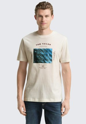 Beige Kurzarm-Baumwoll-T-Shirt mit einem blauen grafischen Quadrat und rotem Text. Kombiniert mit dunkelblauen Jeans, was einen lässigen Look ergibt.