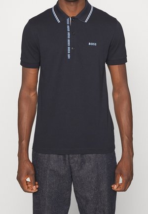 Homme portant un polo bleu marine avec le logo BOSS sur la poitrine et une patte de boutonnage, associé à un jean foncé, debout les bras détendus.