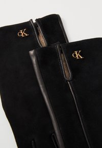 Gants en suede noir avec des accents en cuir, dotés d'une fermeture éclair et d'un détail de logo doré. Texture lisse avec un design ajusté.