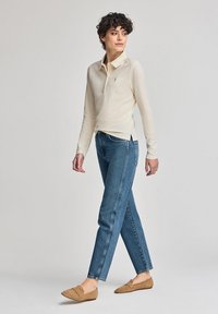 Beige långärmad pikétröja i lätt tyg, med krage och sidslitsar, kombinerad med blå raka jeans och bruna slip-on skor.