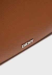 Καφέ δερμάτινη τσάντα με υφή και ασημένιο λογότυπο DKNY σε γεωμετρική γραμματοσειρά. Η επιφάνεια παρουσιάζει λεπτομέρειες με υφή σαν μικρές πέτρες και γυαλιστερό φινίρισμα.