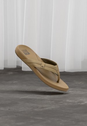 Sandale beige style flip-flop avec détails de couture et semelle texturée, suspendue au-dessus d’un sol en béton gris devant un rideau blanc.