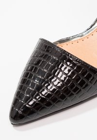 Chaussure noire en cuir verni à bout pointu avec texture crocodile et semelle intérieure beige à détails perforés, photographiée sur un fond blanc.