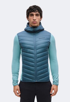 Homme portant un gilet matelassé sans manches bleu avec capuche par-dessus une chemise à manches longues bleu clair, debout devant un fond uni.
