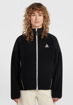 Jong persoon met krullend haar die een zwarte fleecejas met rits draagt met een klein driehoekig logo op de borst en beige broek.